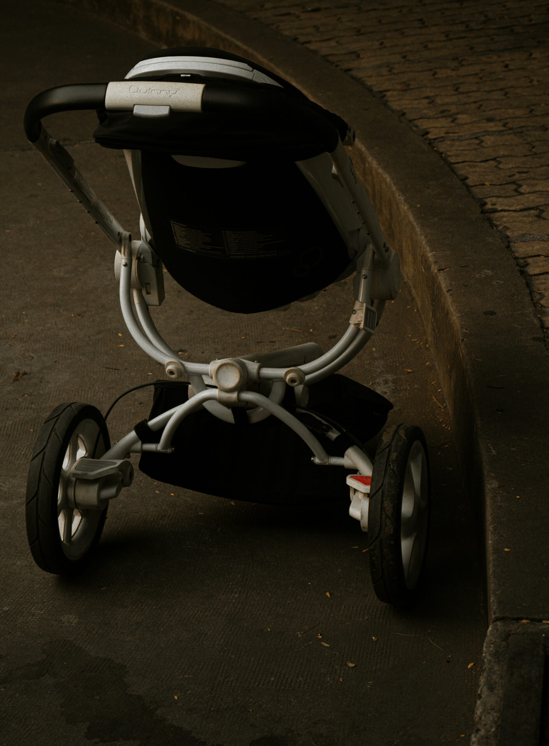 A high-angle, vertical shot of a modern, three-wheeled baby stroller positioned on a paved path. The stroller is viewed from behind, showing its silver frame, black seating area, and large, rugged wheels. The lighting is moody and focused, with a textured stone curb running along the right side of the frame.